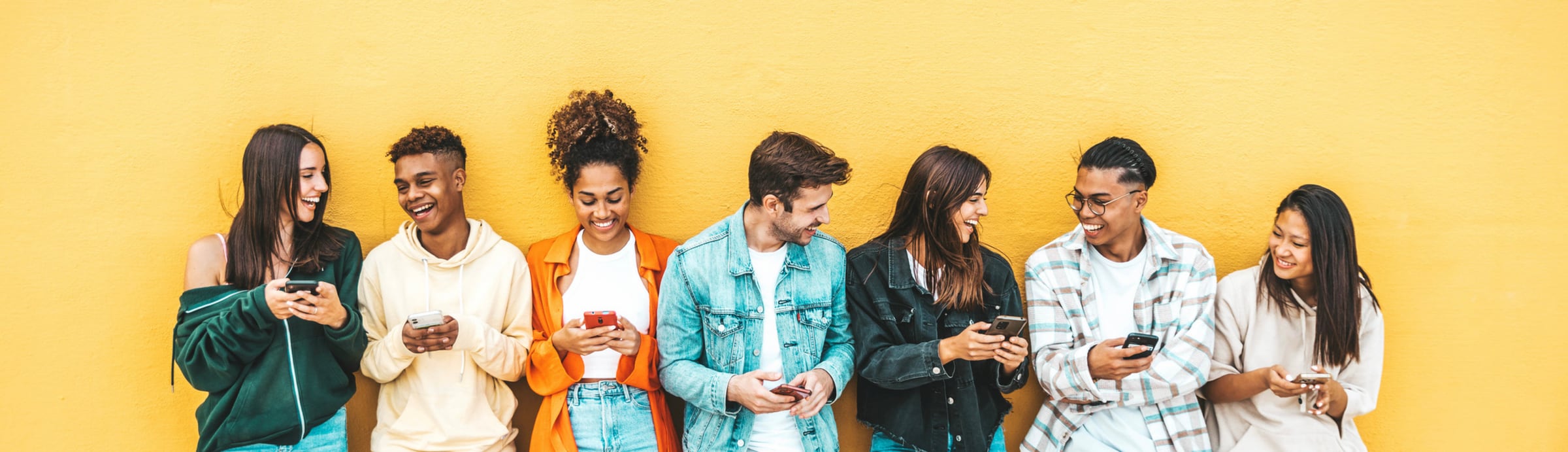 Seven colleagues leaning against a yellow wall, each looking at their phone and smiling
